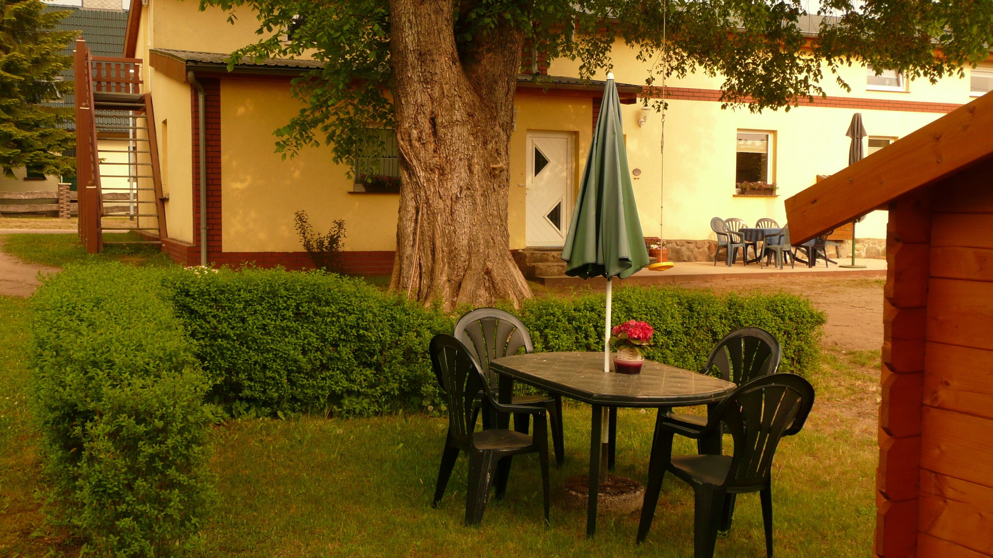 Un espacio al aire libre con mesa y sillas, un árbol y un edificio al fondo.