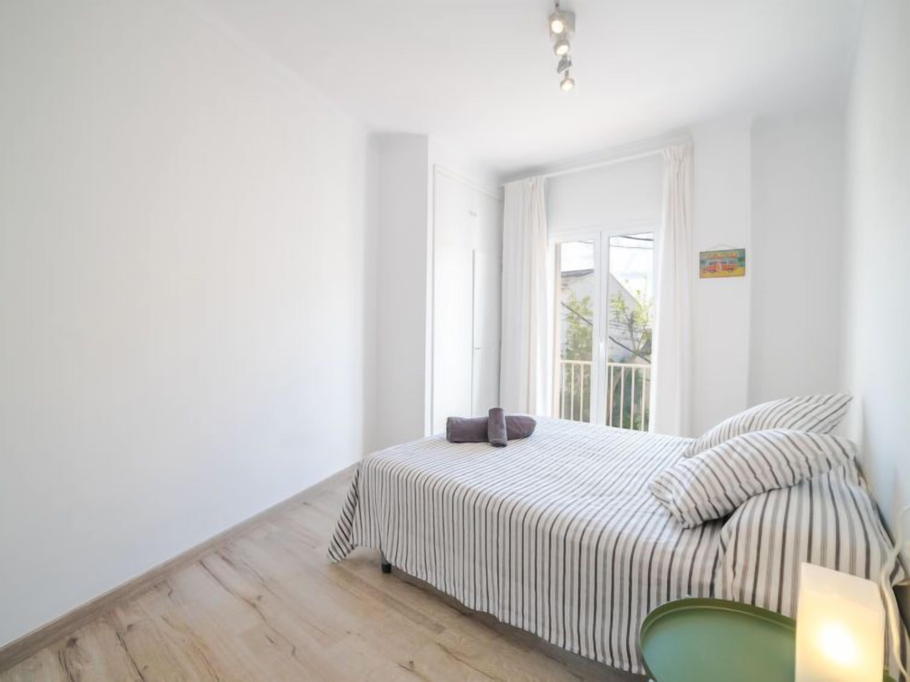 A bright, minimalist bedroom with a large window, a striped bed, and a green chair.