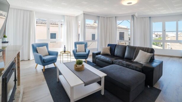 Una sala moderna con un sofá de cuero negro, dos sillones azules, una mesa de centro blanca y un ventanal con vistas a edificios.