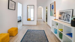 Un pasillo con un espejo, cuadros enmarcados, un reposapiés amarillo, una alfombra azul y una consola blanca con estantes.