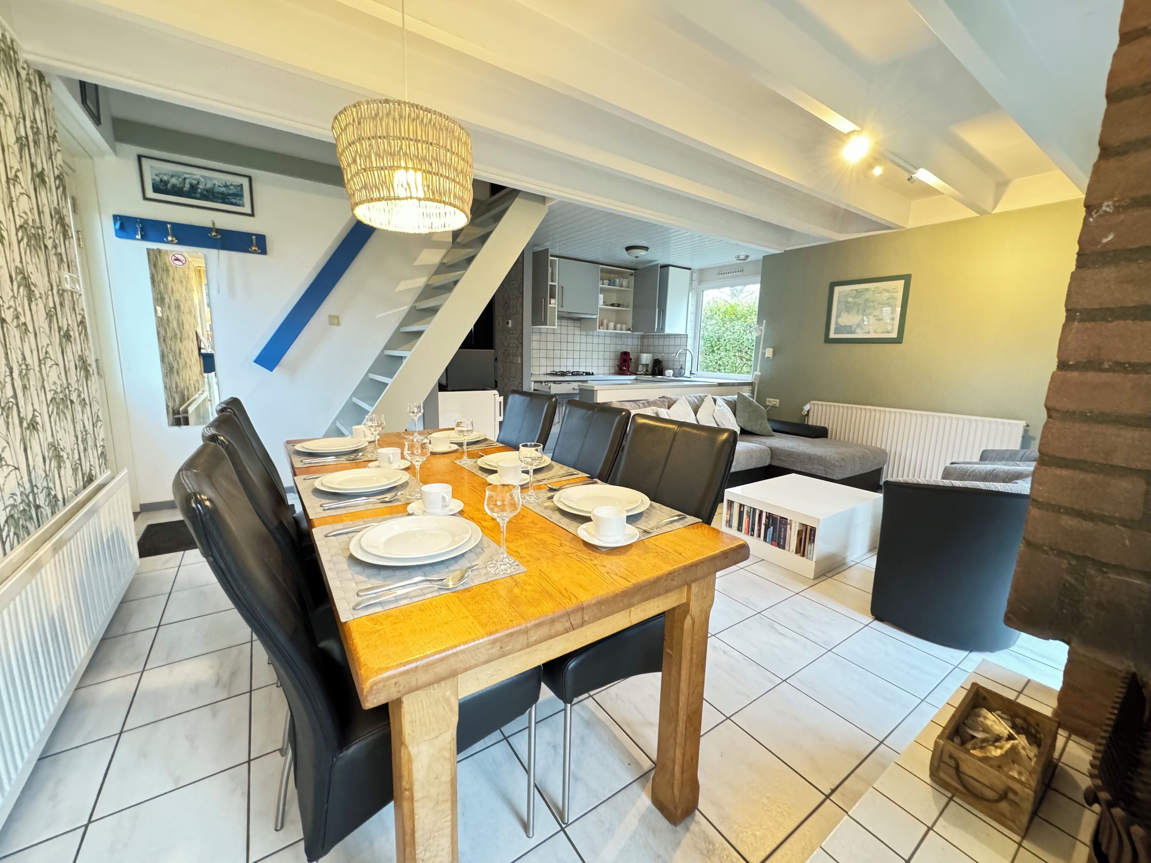 Un comedor con una mesa de madera preparada para cuatro personas, una escalera que conduce a una cocina y una sala de estar con un sofá y una chimenea.