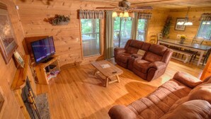 A cozy cabin interior with a wooden floor, leather sofa, a television, and a dining area. - Fox Ridge- Perfect cabin nestled in the woods with a large stacked rock firepit (Sautee Nacoochee)