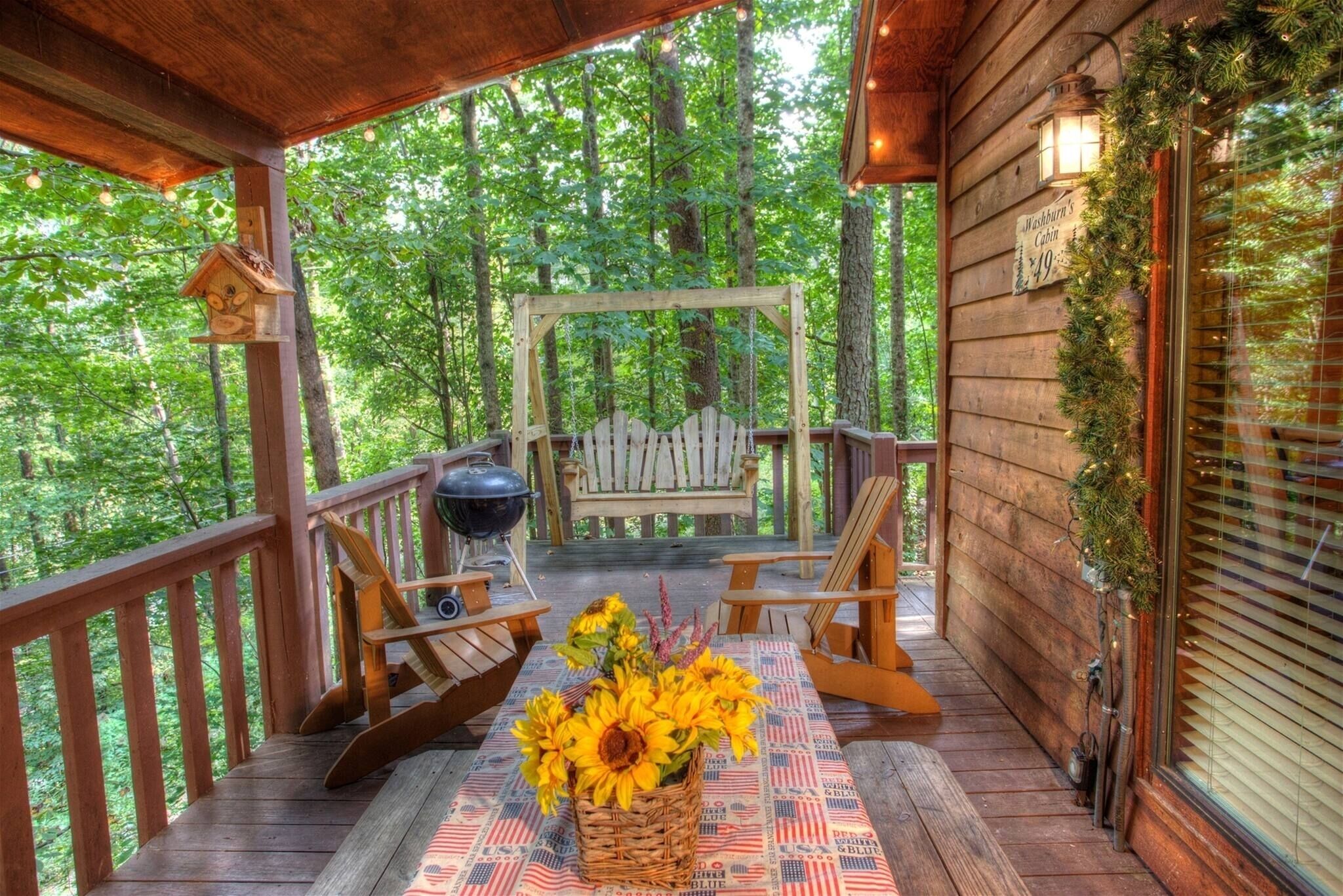 Eine Holzterrasse mit Grill, zwei Adirondack-Stühlen, einem Tisch mit Sonnenblumen-Dekoration und einem Schild, das auf eine Kabinenmiete hinweist.