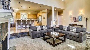 A spacious living room with a gray sectional sofa, a coffee table, and a fireplace. A kitchen is visible in the background with wooden cabinets and a dining area.