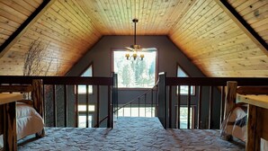 Ein Schlafzimmer im Loft-Stil mit schrägem Dach, Holzbalken, einem Bett, einer Treppe und einem Fenster mit Blick auf Bäume.