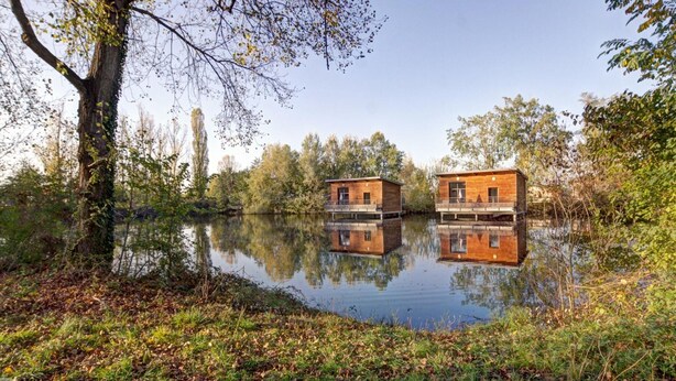 Deux maisons en bois, construites sur des pontons, flottent sur un lac paisible, entouré d’arbres.