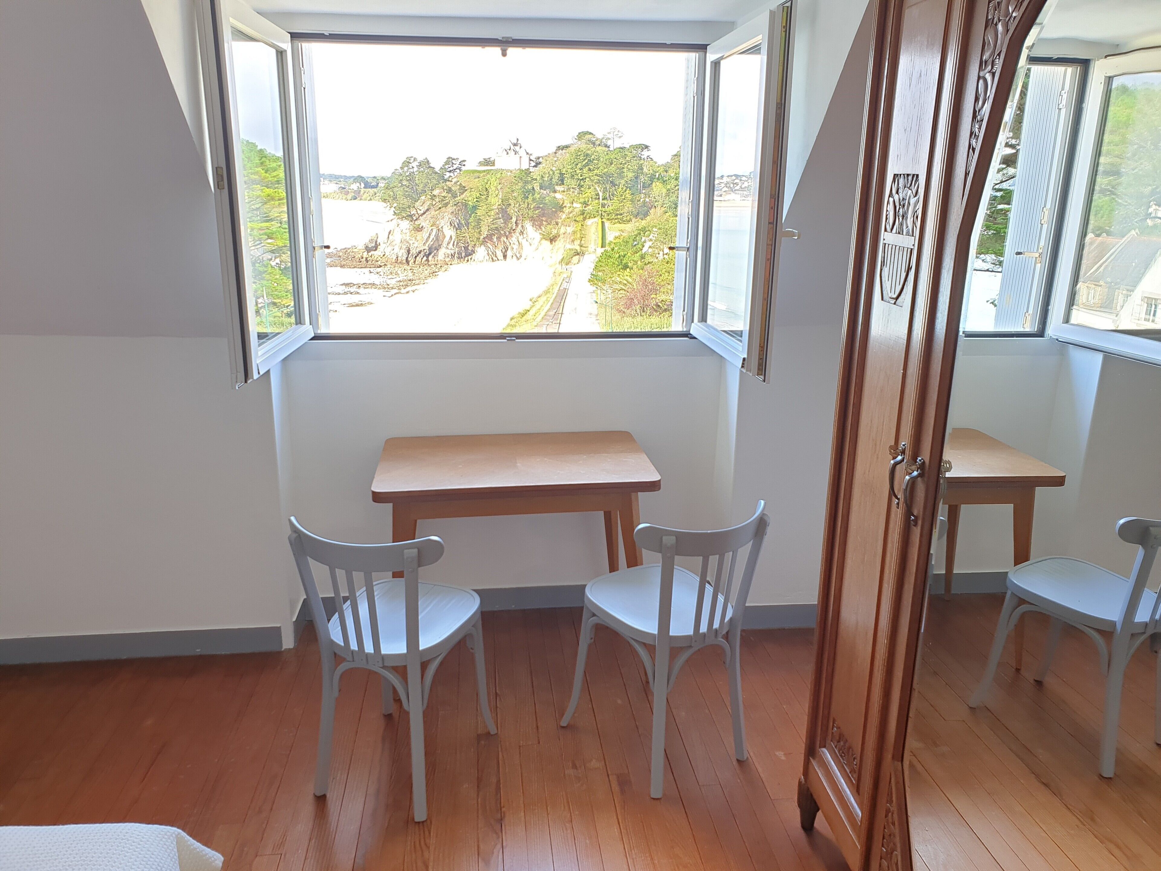 Ein Zimmer mit Holzboden, einem Holzbüro und zwei weißen Stühlen. Es gibt ein Fenster mit Blick auf einen Strand und Grünflächen.
