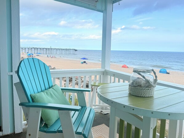 A beach view from a covered porch with a chair and table, a pier in the distance, and beach umbrellas. - Our BIG BEAUTIFUL BEACH is back Amazing Oceanfront Condo w/ Breathtaking VIEWS (Carolina Beach)