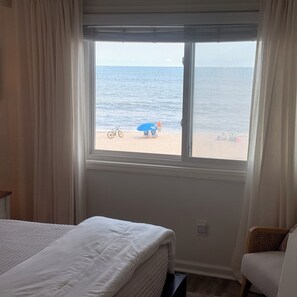Ein Schlafzimmer mit großem Fenster und Blick auf einen Strand, einem Bett und einem Sessel.