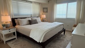 Une chambre avec un grand lit, deux tables de chevet avec des lampes, une plante sur l’une des tables de chevet, et une vue sur l’extérieur par la fenêtre.