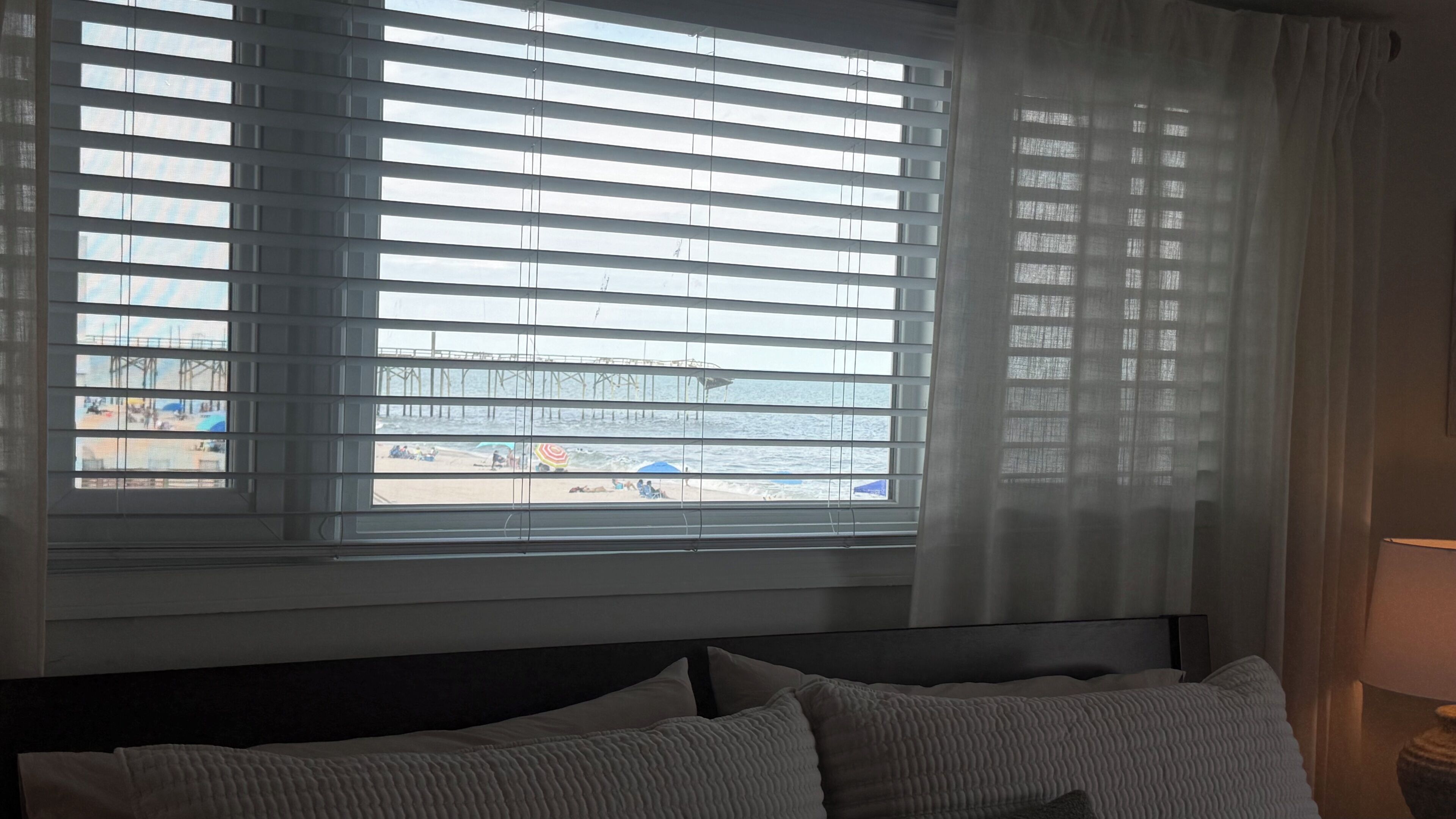 Ein Hotelzimmer mit Bett, einem Fenster mit Jalousien und Blick auf Strand und Steg.