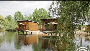 Two wooden cabins with outdoor seating, connected by a bridge over a calm lake, surrounded by trees. - Unique cabin by a pond, pets allowed, private terrace (Veauchette)