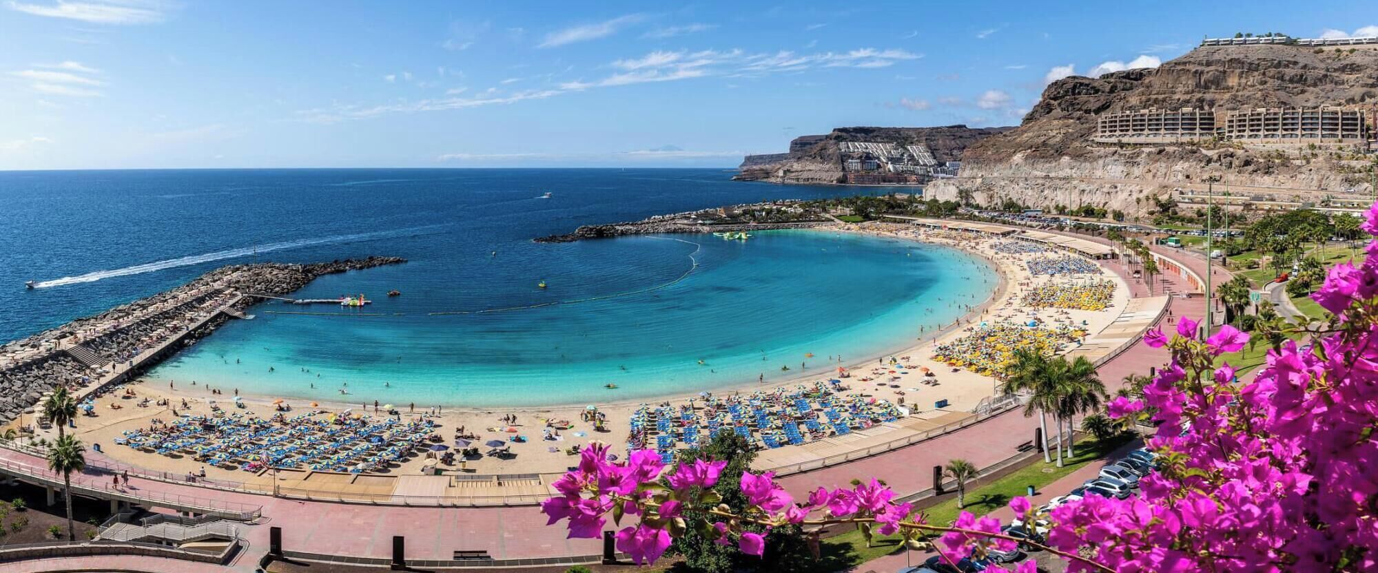 A coastal resort with a large sandy beach, a rocky breakwater, and a mountainous backdrop.