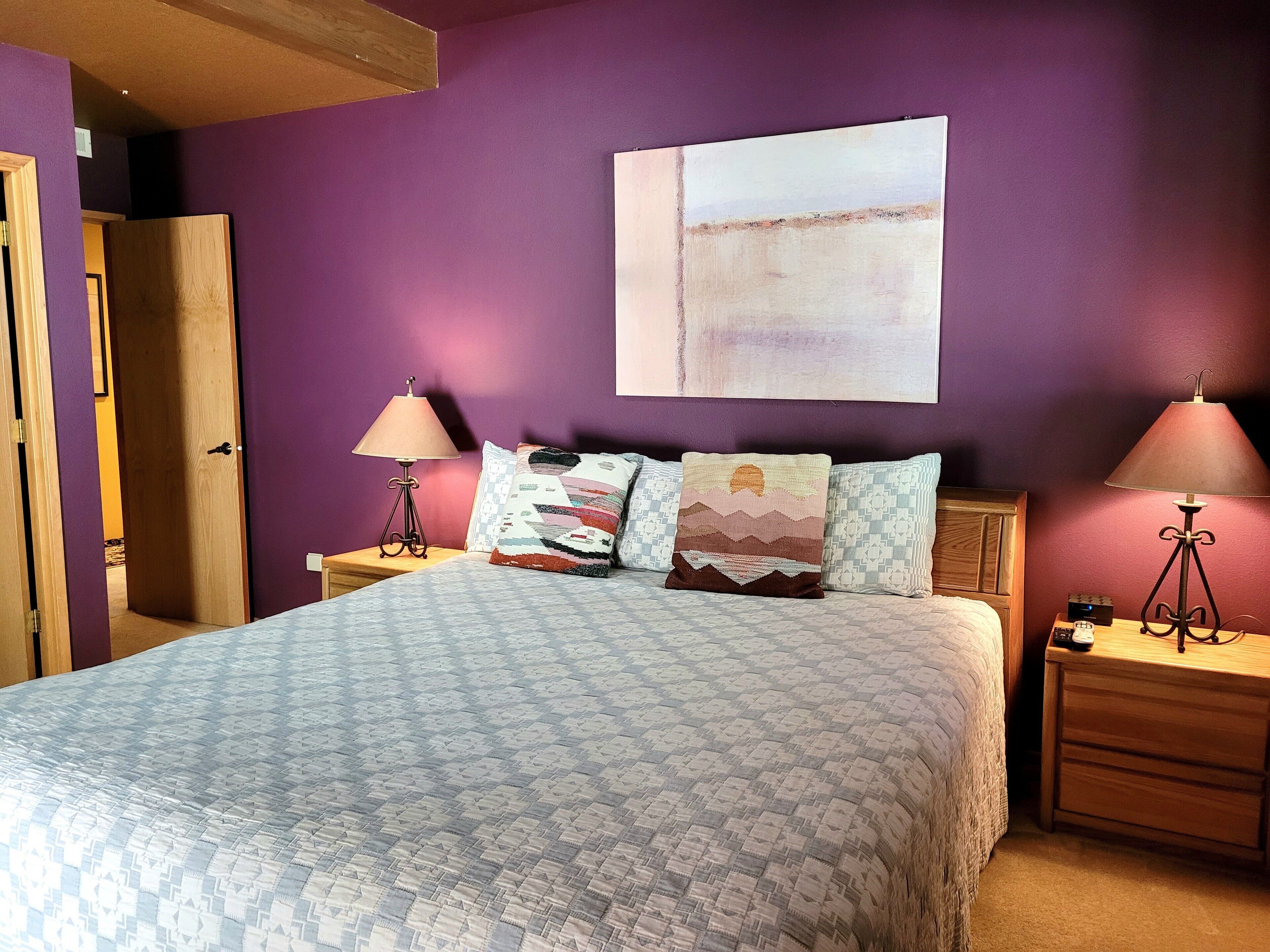 A bedroom with a bed, two nightstands, a wall-mounted lamp, a door, and a painting.
