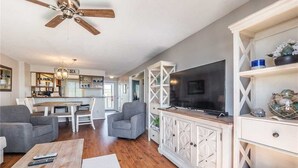 A spacious living room with a large flat-screen TV, grey sofas, wooden flooring, and a ceiling fan. - Take It E-Sea | Oceanfront + Community Pool! (Kure Beach)