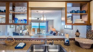 A kitchen with a double sink, wooden shelves with glassware, a view of the living area, and a sign that says 'My heart is on the shore.' - Take It E-Sea | Oceanfront + Community Pool! (Kure Beach)