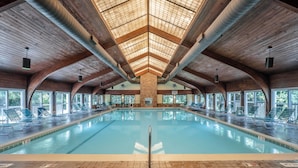 A large indoor swimming pool with a high ceiling and wooden beams, surrounded by lounge chairs and large windows. - Take It E-Sea | Oceanfront + Community Pool! (Kure Beach)