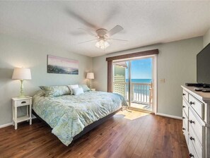 A bedroom with a bed, nightstands, a dresser, a ceiling fan, and a sliding glass door leading to a balcony. - Take It E-Sea | Oceanfront + Community Pool! (Kure Beach)