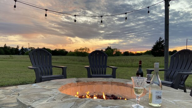 A stone fire pit with chairs around it, a bottle of wine, and a glass on a table.