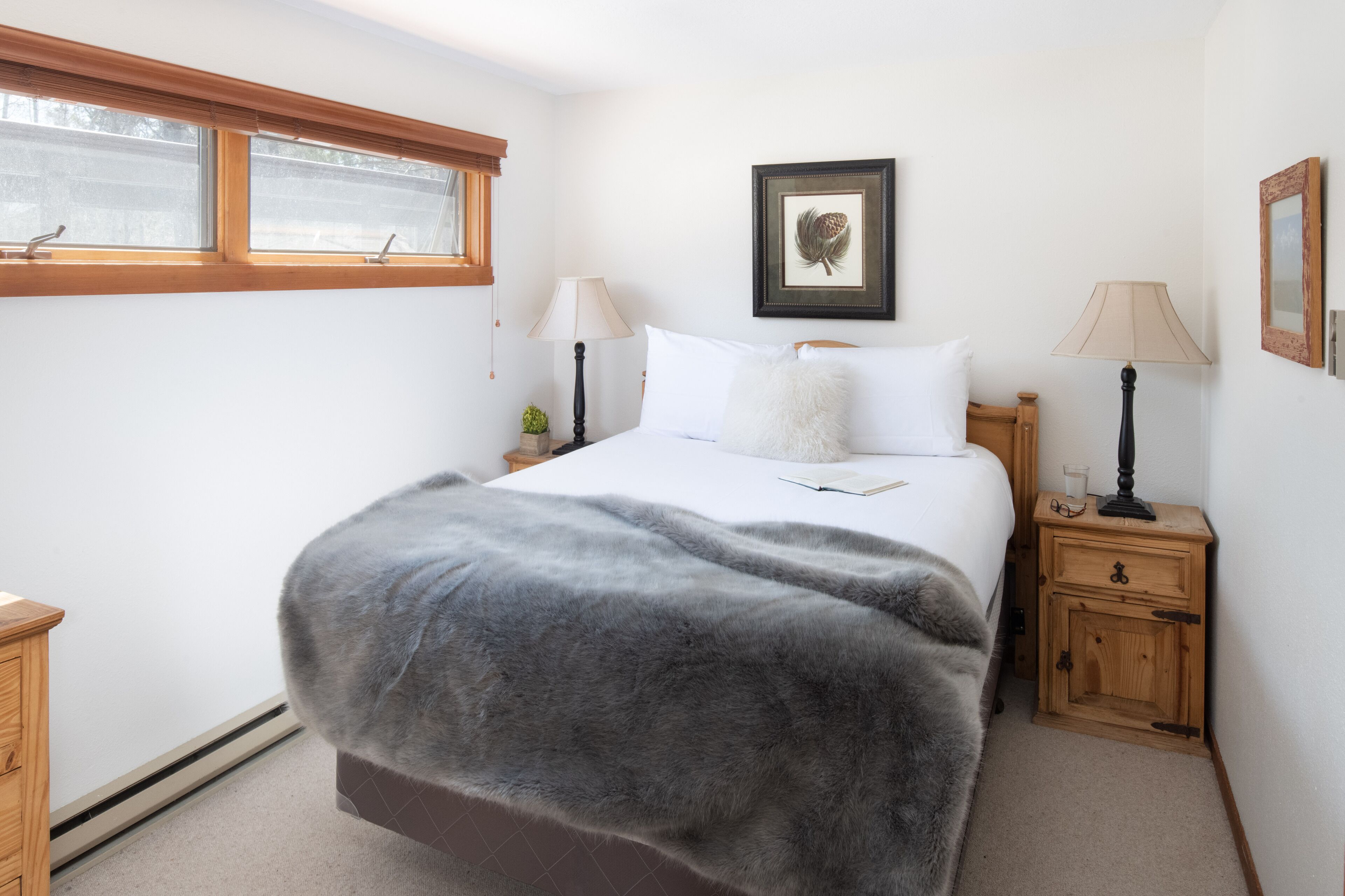 A bedroom with a large bed, two bedside tables with lamps, a framed picture on the wall, and a window with blinds.