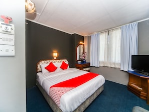 Une chambre d’hôtel avec un lit, une télévision, une coiffeuse avec un miroir et une fenêtre avec des rideaux.