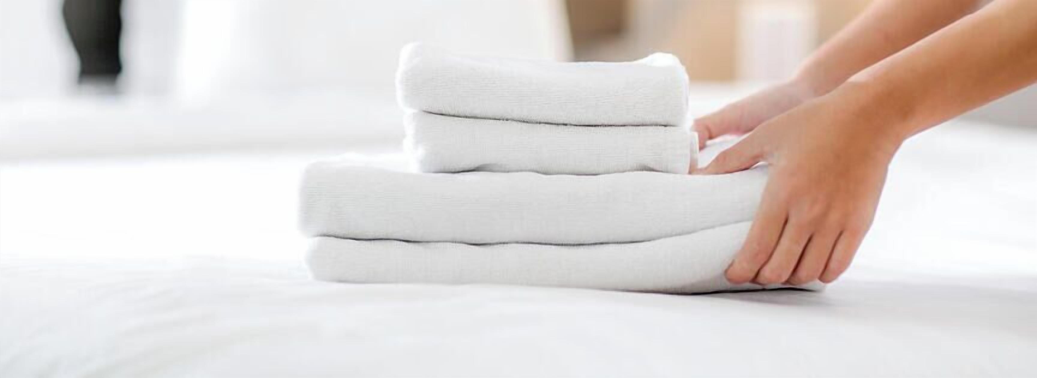 A person folding a stack of white towels on a bed.