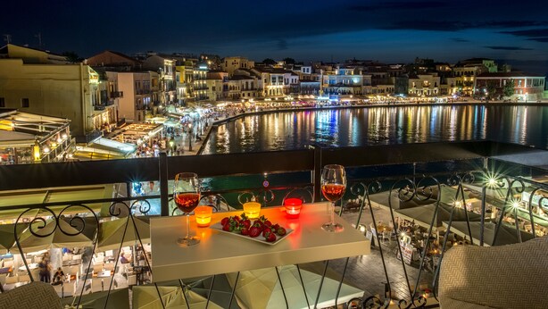 Une terrasse avec une table dressée pour deux personnes, donnant sur un front de mer bordé de bâtiments et dont l’eau reflète la lumière.