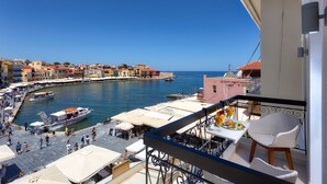 Un balcon avec une chaise en osier blanche et une table sur laquelle se trouve une assiette de nourriture, donnant sur un port où l’on peut voir des bateaux et des bâtiments aux couleurs vives.