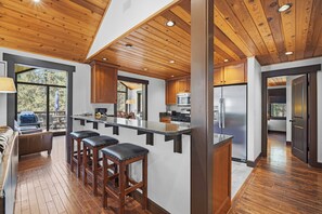 A modern kitchen with wooden ceiling, bar stools, and a view to the outside.