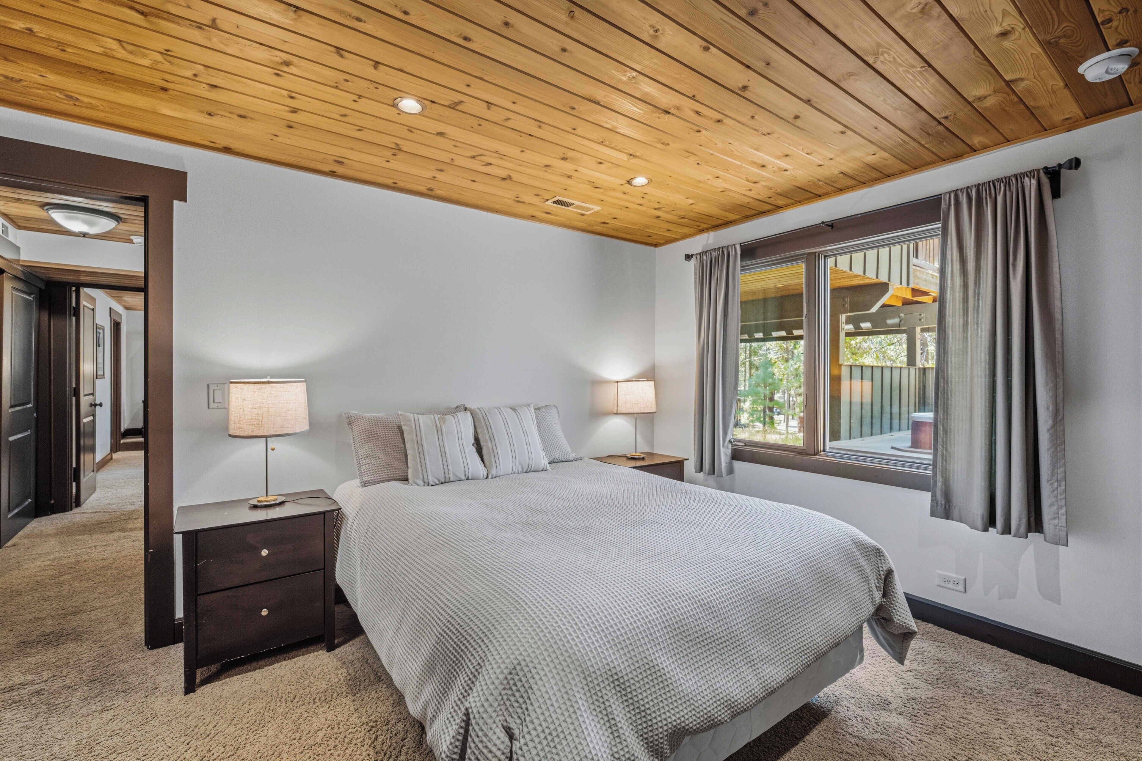 Une chambre à coucher avec un plafond en bois, un lit recouvert d’une couette grise, deux lampes de chevet et une fenêtre avec des rideaux.