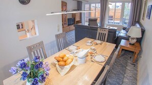Ein Essbereich mit einem Holztisch, gedeckt für das Frühstück, einer Blumenvase und Blick auf die Küche und das Wohnzimmer.