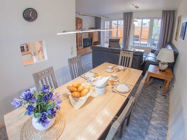 A dining area with a wooden table set for breakfast, a vase of flowers, and a view of the kitchen and living area.