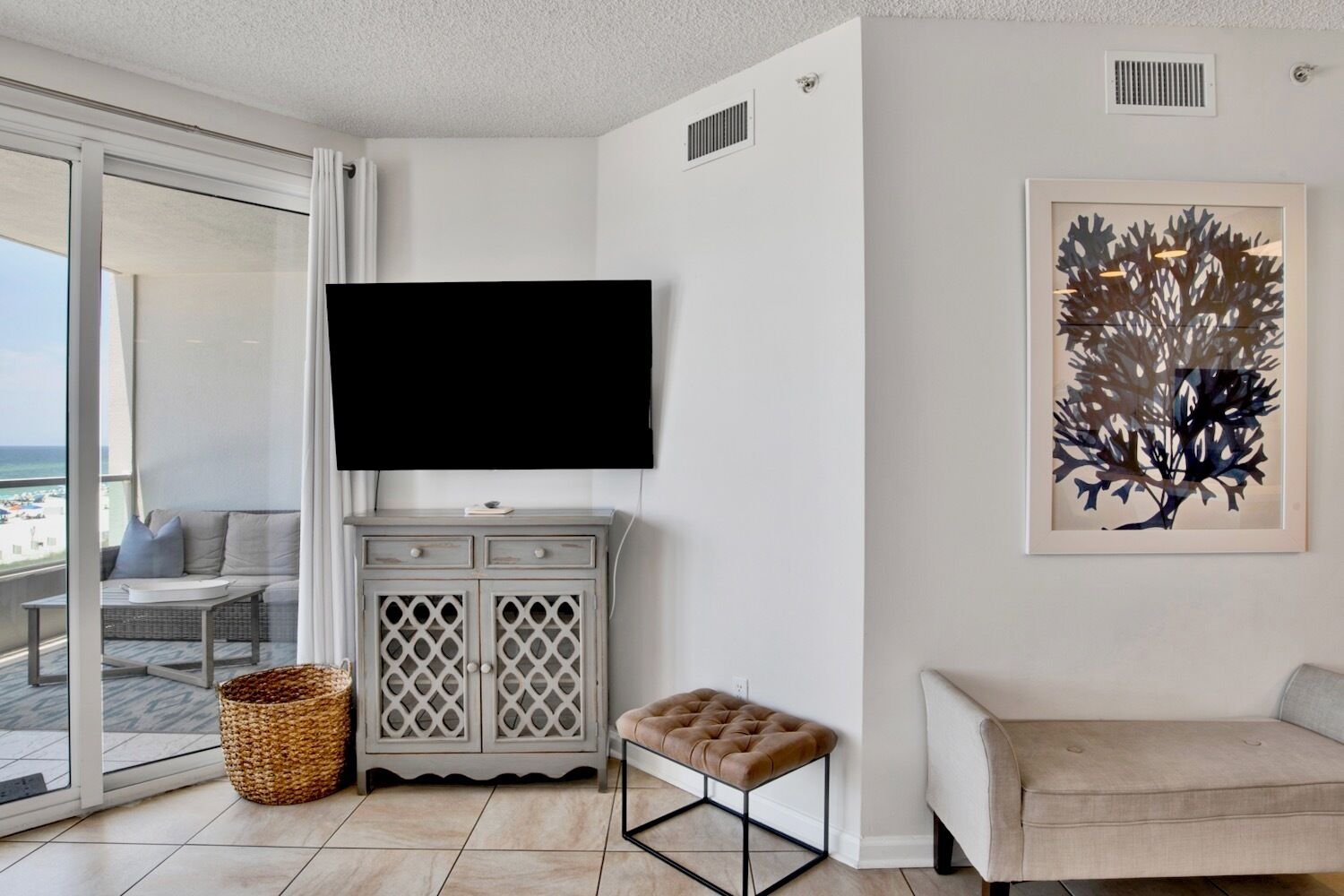 A modern living room with a large flat-screen TV mounted on the wall, a grey cabinet with drawers, a cushioned bench, and a wicker basket on the floor.