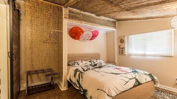 Quarto com cama, janela com persianas, cortina de bambu e parede com pintura.