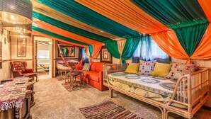 Una habitación acogedora y bien iluminada con una cama, una silla y una pequeña mesa. Hay cortinas coloridas y una alfombra estampada en el suelo.