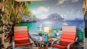 Habitación con un mural de una playa tropical, dos sillones rojos acolchados, una mesita redonda con una lámpara y una planta en maceta.