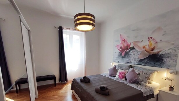 A bedroom with a bed, a bench, a painting of flowers, and a hanging light fixture. - Orchidea Apartment Split in the center (Split)