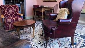 Ein Wohnzimmer mit einem dunkelroten Ledersessel, einem gemusterten Sessel, einem Holztisch, einem Esstisch mit Stühlen und einer Küche im Hintergrund.