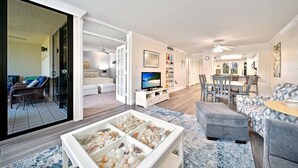 A spacious living room with a large glass door leading to a balcony, a dining area with a wooden table and chairs, and a kitchen in the background. - Loggerhead Cay #322 - Sanibel Island Beach View (Sanibel)