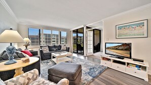 A modern living room with a sofa, ottoman, coffee table, and TV stand. There is a large window with blinds, a door leading to a balcony, and a framed picture on the wall. - Loggerhead Cay #322 - Sanibel Island Beach View (Sanibel)