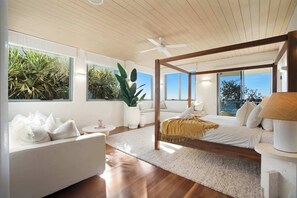 Una camera da letto moderna con soffitto in legno, un letto a baldacchino, un divano bianco e ampie finestre con piante tropicali.
