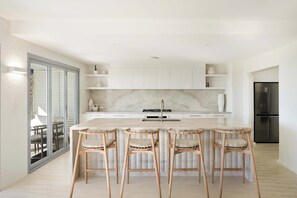 Une cuisine moderne dotée d’un comptoir en marbre, de quatre tabourets de bar en osier, d’un réfrigérateur en acier inoxydable et d’une porte-fenêtre coulissante donnant sur un espace extérieur.