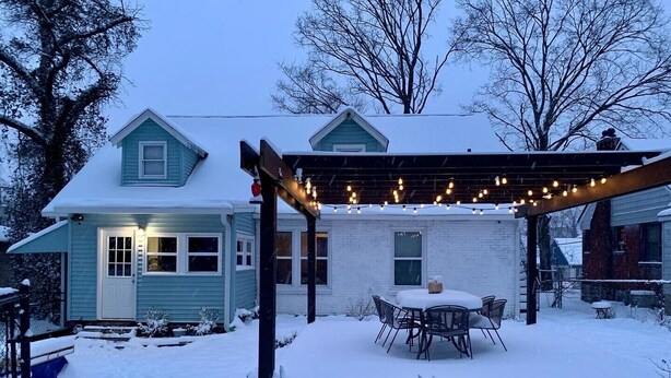 A snow-covered backyard with a patio area featuring a table and chairs, string lights, and a pergola.