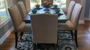A dining area with a table set for four, surrounded by upholstered chairs, a patterned rug, and a window with a view of greenery.
