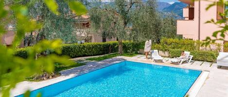 Una piscina rectangular con agua azul cristalina, rodeada de vegetación y un edificio con balcones.