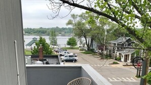 Un balcón con una mesa y sillas rojas, una pequeña planta en maceta y vista a una calle y un lago.