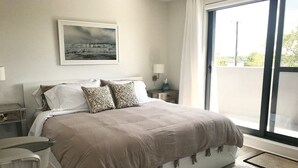 Un dormitorio con una cama grande, una mesita de noche, una lámpara, una ventana con cortinas y un cuadro enmarcado en la pared.