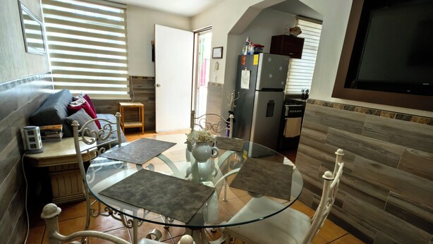 A compact living space with a dining area featuring a glass-top table and metal chairs, a kitchen with a built-in refrigerator and a TV mounted on the wall, and a door leading to an adjacent room. - Charming little house in Tlaquepaque (San Juan)