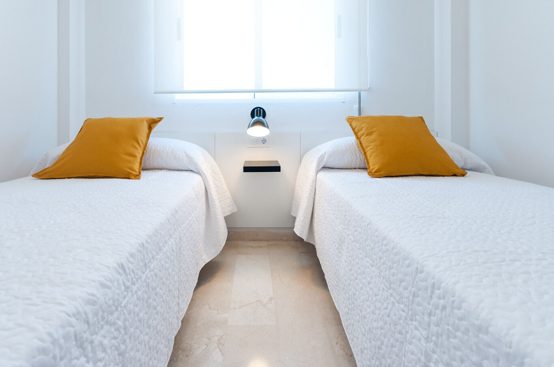 Ein Zimmer mit zwei Betten, jedes mit einer weißen Bettdecke und einem senfgelben Kissen. Dazwischen steht ein kleiner Nachttisch mit einer Lampe.