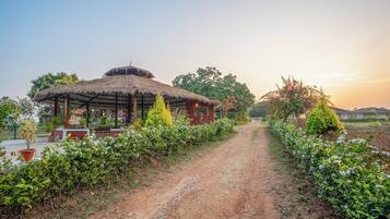 A rustic thatched-roof structure with a dirt path leading to it, surrounded by greenery and trees.
